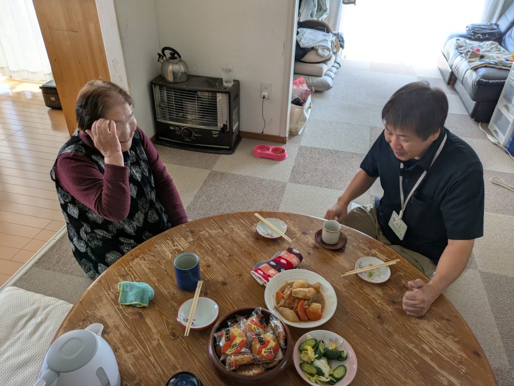 Caregiver sharing a homemade meal and chatting with an elderly woman during a home visit in Sendai, Japan