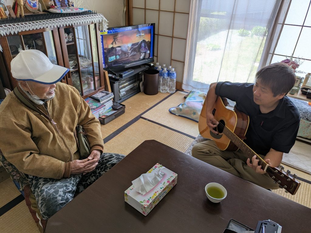 A caregiver playing music for an elderly man during an elderly care and monitoring service visit in Sendai.