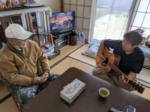 A caregiver playing music for an elderly man during an elderly care and monitoring service visit in Sendai.